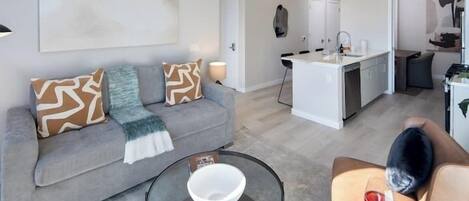 Una sala de estar moderna con un sofá gris, una mesa de centro de vidrio y un comedor con una cocina blanca al fondo.