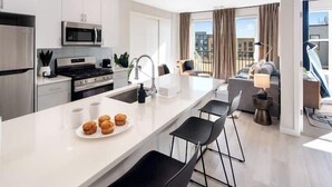 A modern kitchen with white cabinetry, a central island with bar stools, and a view of the city through large windows. - Luxury Furnished Apartments by Hyatus Downtown at Yale (New Haven)