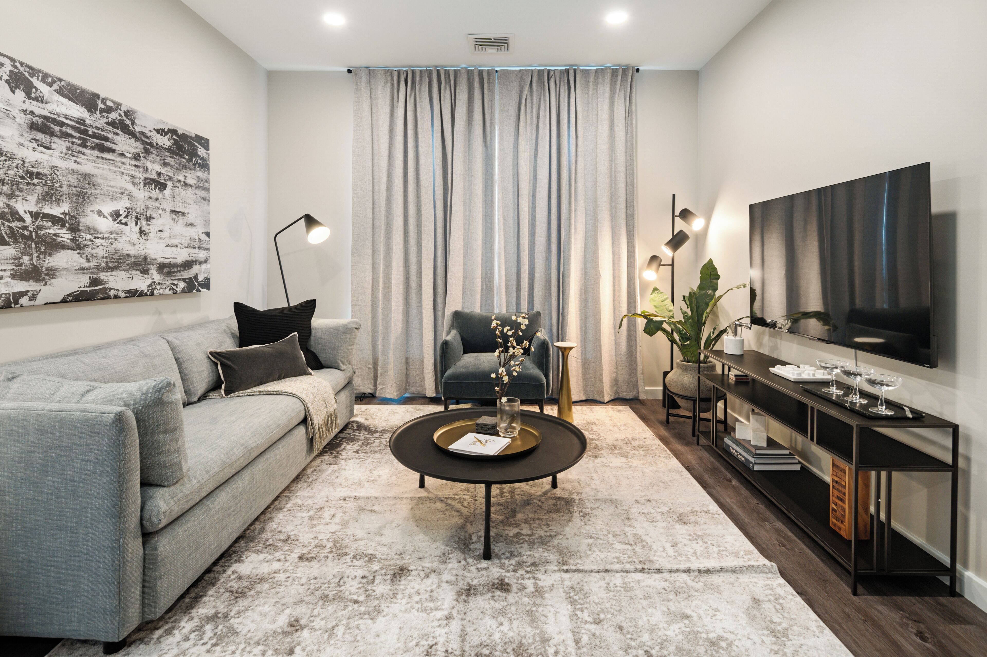 A modern living room with a grey sofa, a round coffee table, a blue armchair, a TV, and a large window with curtains.