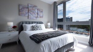 A modern bedroom with a large bed, two bedside tables, a window with a city view, and abstract wall art. - Luxury Furnished Apartments by Hyatus Downtown at Yale (New Haven)