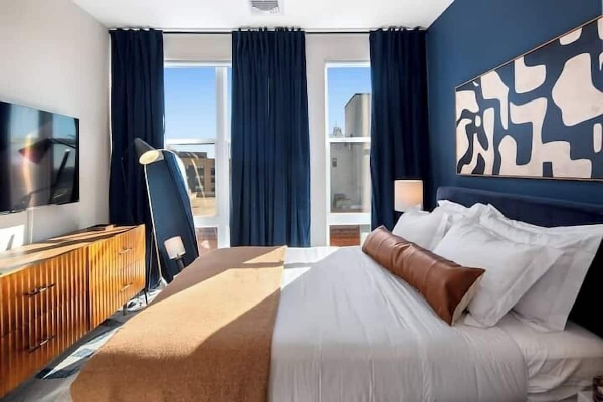 A hotel room with a large bed, a TV, a wooden dresser, and a view of the city through large windows.