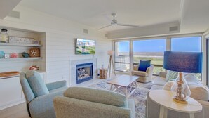 Una sala de estar con chimenea, ventilador de techo, un ventanal con vistas y un televisor montado en la pared.