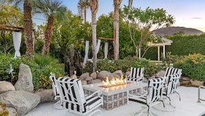 Un patio al aire libre con una chimenea, sillas blancas a rayas y una pérgola con telas colgantes.