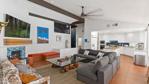 Una sala de estar moderna con un sofá gris, un banco rojo y una mesa de centro de vidrio. Se aprecia una cocina al fondo con armarios blancos y un salpicadero azul.