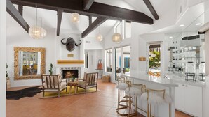 Amplio salón con chimenea, vigas de madera y un adorno de cabeza de toro. Hay un comedor con sillas amarillas y un espejo grande. La cocina tiene una encimera blanca y cristalería.