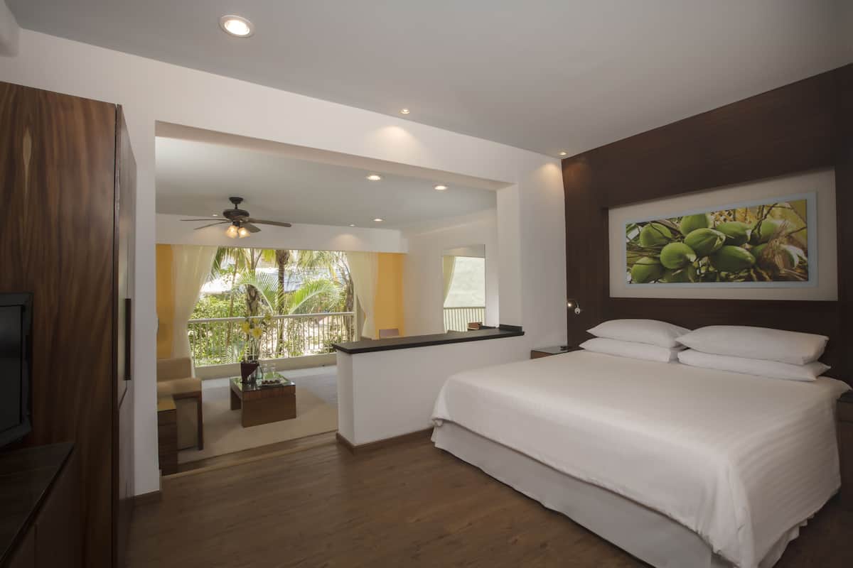A bedroom with a bed, a ceiling fan, a balcony with a view of greenery, and a painting on the wall.