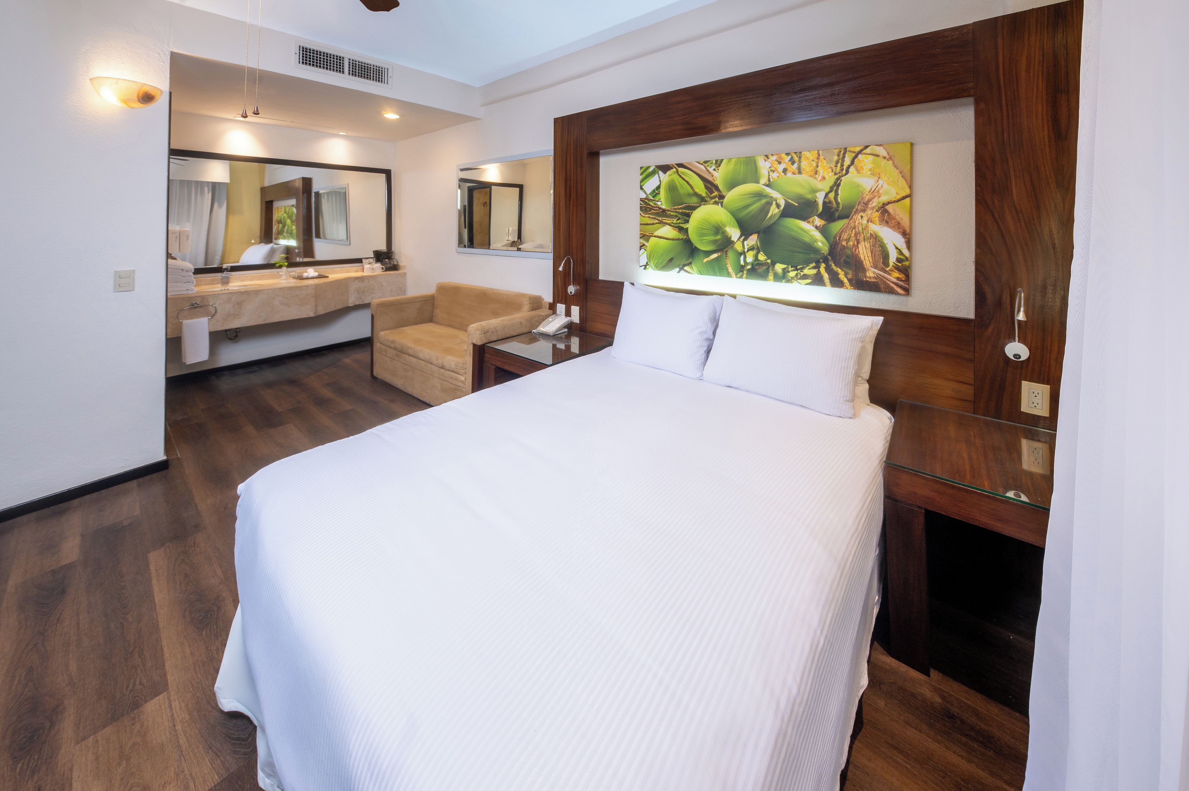 A hotel room with a large bed, a sitting area with a chair, a vanity with a mirror, and a wall-mounted picture.