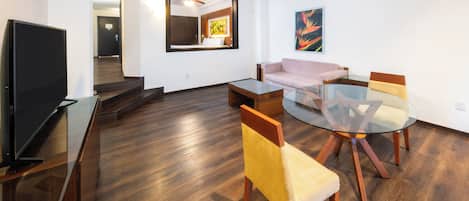 A modern living room with a flat-screen TV, a glass-top dining table with chairs, a sofa, and a ceiling fan.