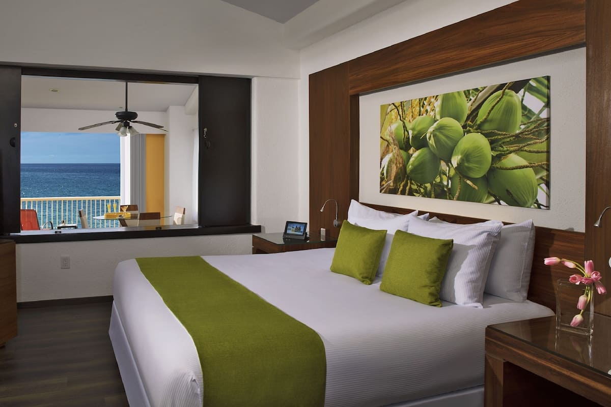 A hotel room with a bed, a TV, a balcony with a view of the ocean, and a painting on the wall.