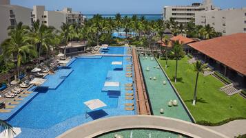 Una amplia piscina con múltiples carriles, rodeada de sillones de descanso y palmeras, con vista al océano y edificios de hotel al fondo.