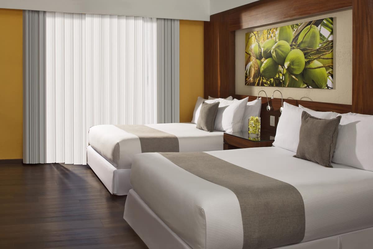 A hotel room with two beds, a large window with curtains, a wooden headboard, and a painting of coconuts.