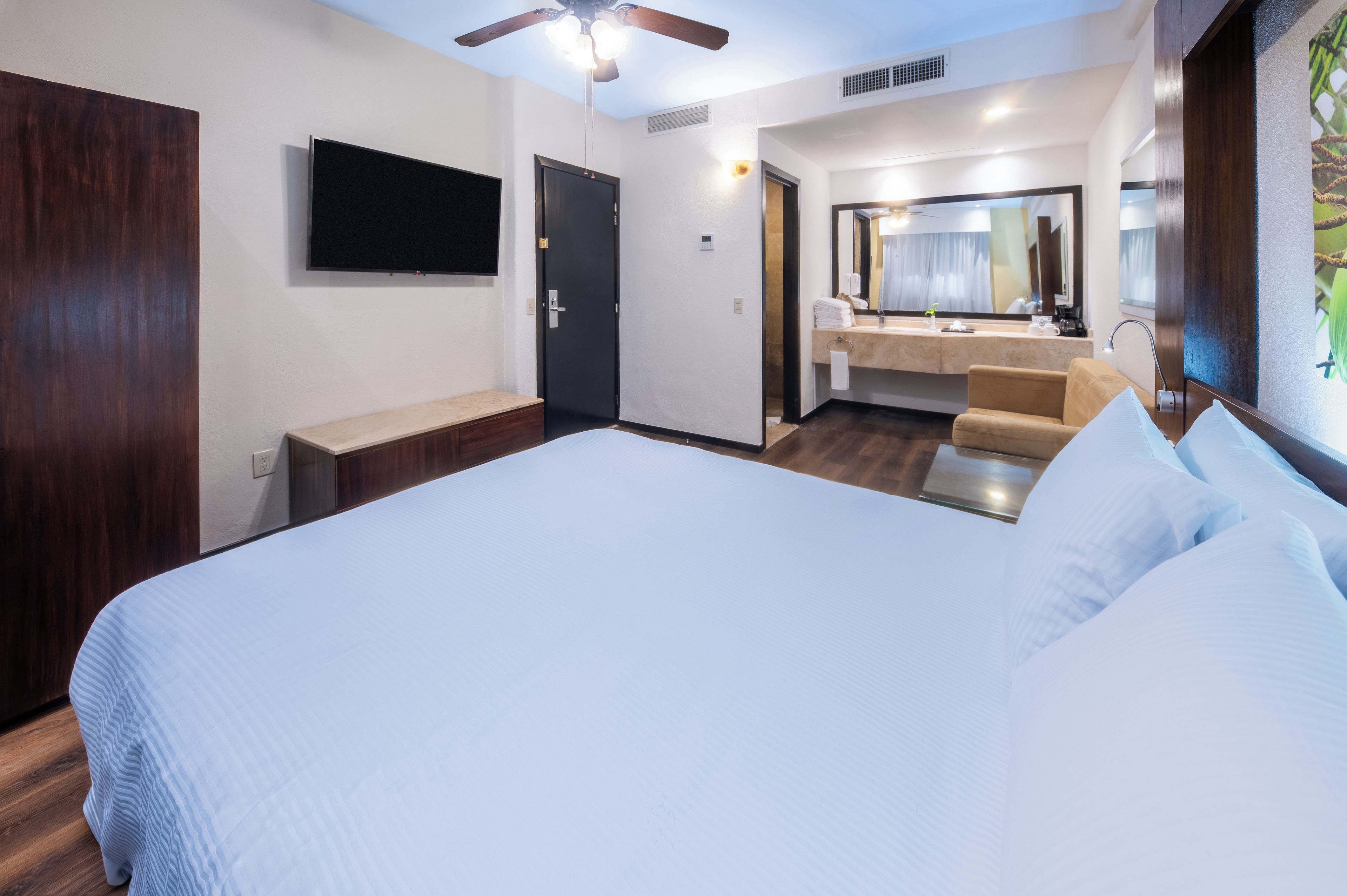 A hotel room with a large bed, a television mounted on the wall, a bathroom with a mirror and sink, and a ceiling fan.