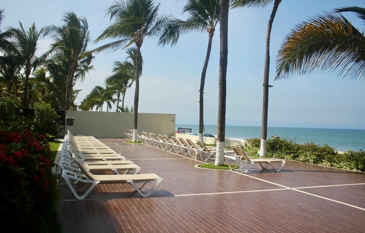 A sunlit deck with lounge chairs, palm trees, and a view of the ocean.