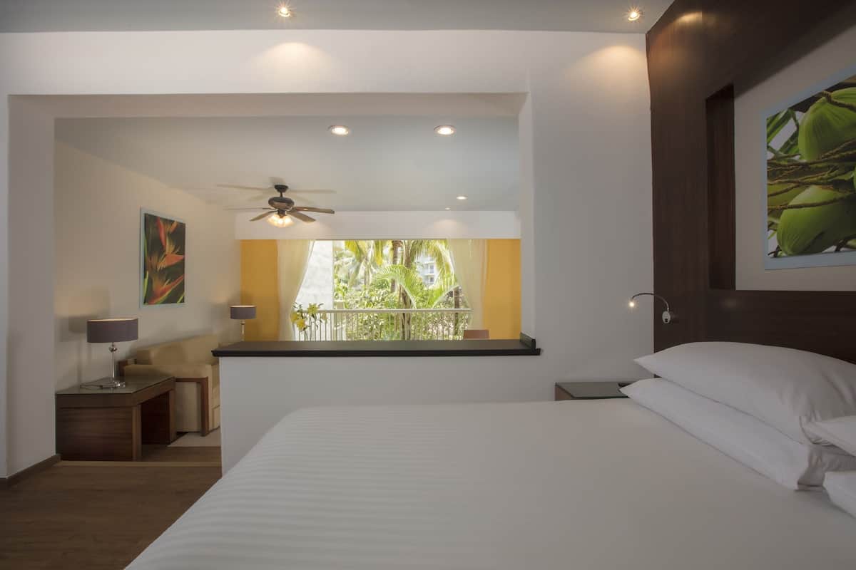 A hotel room with a bed, a desk, a chair, a ceiling fan, and a window with a view of greenery.