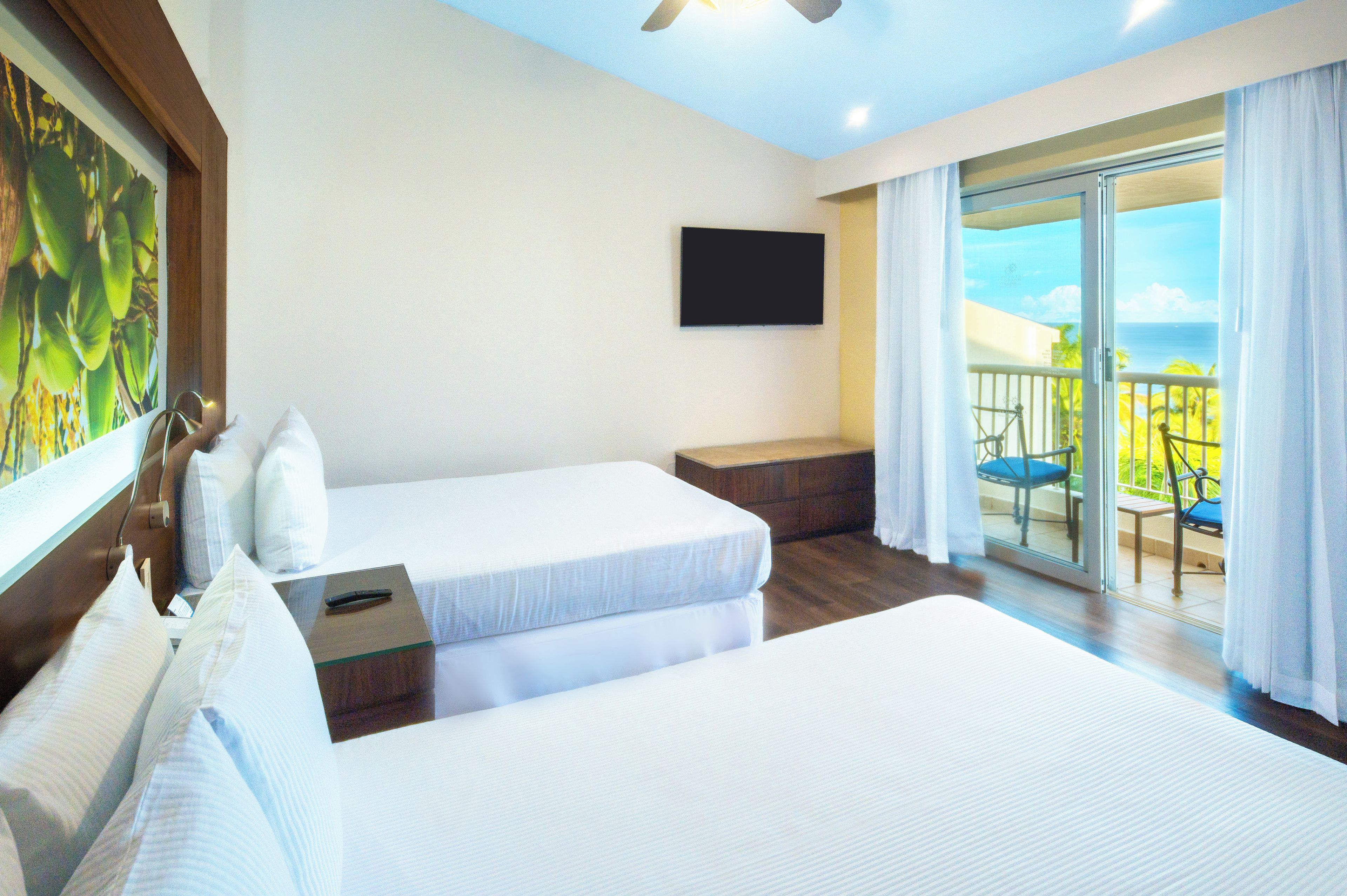 Habitación de hotel con dos camas, un televisor de pantalla plana, un balcón con vista al océano y un cuadro en la pared.