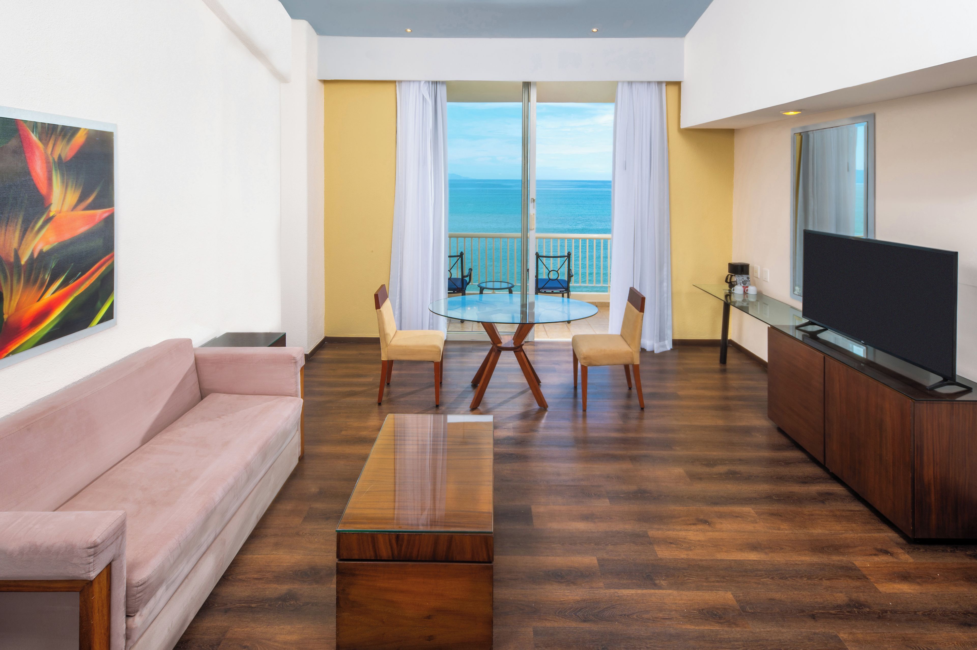 A modern living room with a sofa, a coffee table, a TV, and a balcony with a view of the sea.