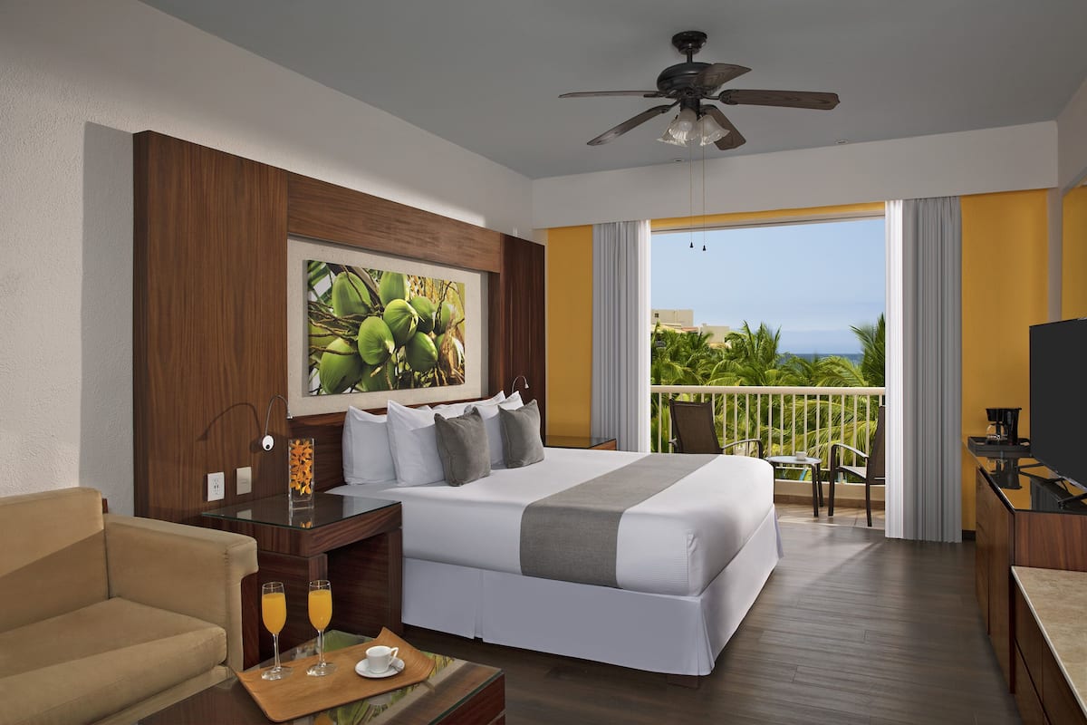 A hotel room with a large bed, a desk with a computer, a TV, a balcony with a view of the ocean, and a ceiling fan.