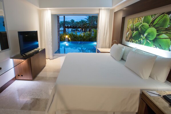 Una habitación de hotel con una cama grande, televisión, escritorio con computadora y vista a una piscina y palmeras.