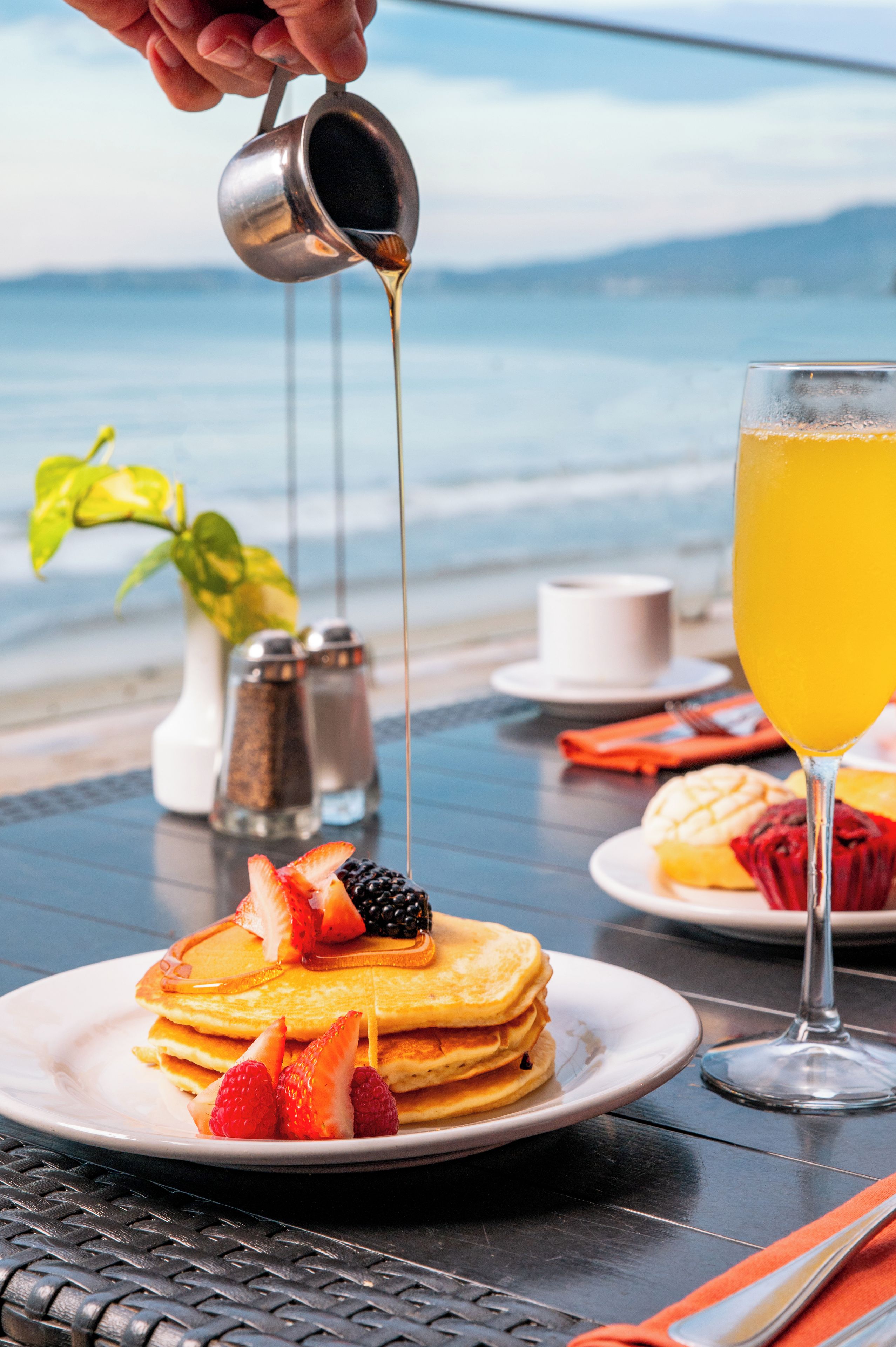 A breakfast setting with pancakes, strawberries, blackberries, and a glass of orange juice on a table with a view of the sea.