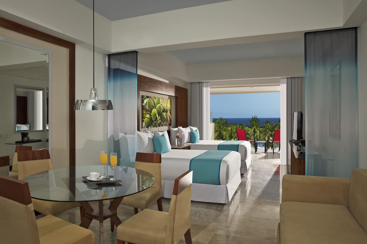 A hotel room with a glass dining table, two beds with teal bedding, a TV, and a view of the ocean.