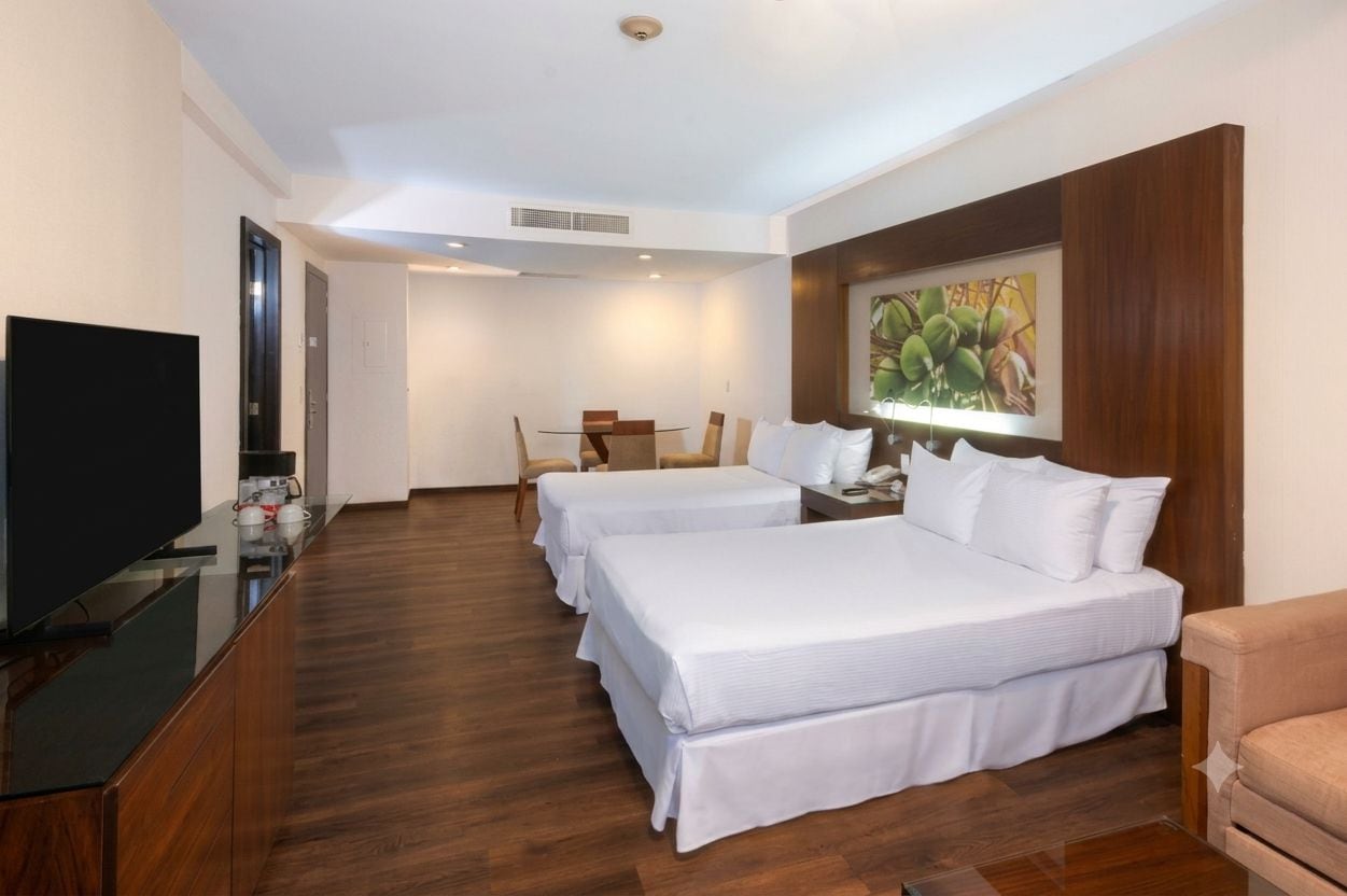 A hotel room with two beds, a television, a dining table with chairs, and a painting on the wall.