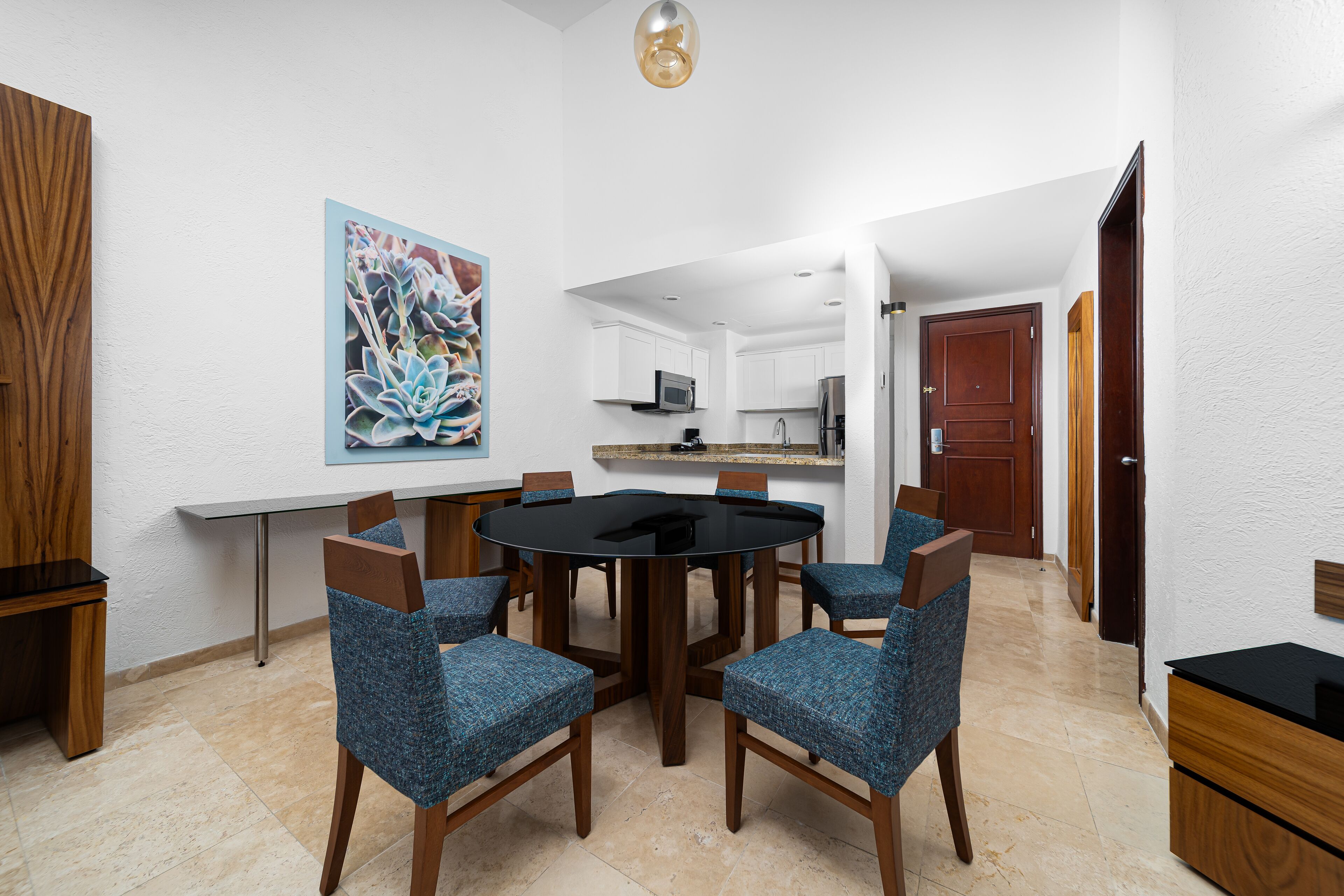 Un comedor con una mesa redonda y sillas tapizadas en azul, un cuadro en la pared y una cocina al fondo.