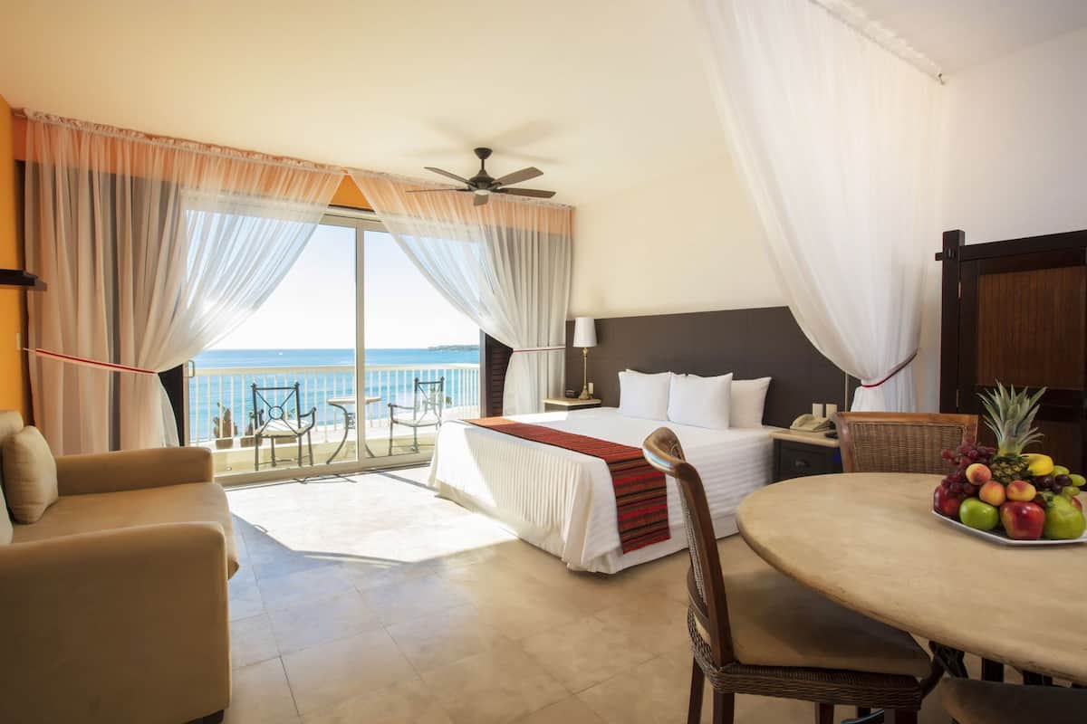 A hotel room with a bed, a dining table with fruit, a chair, a sofa, a balcony with a view of the sea, and a ceiling fan.