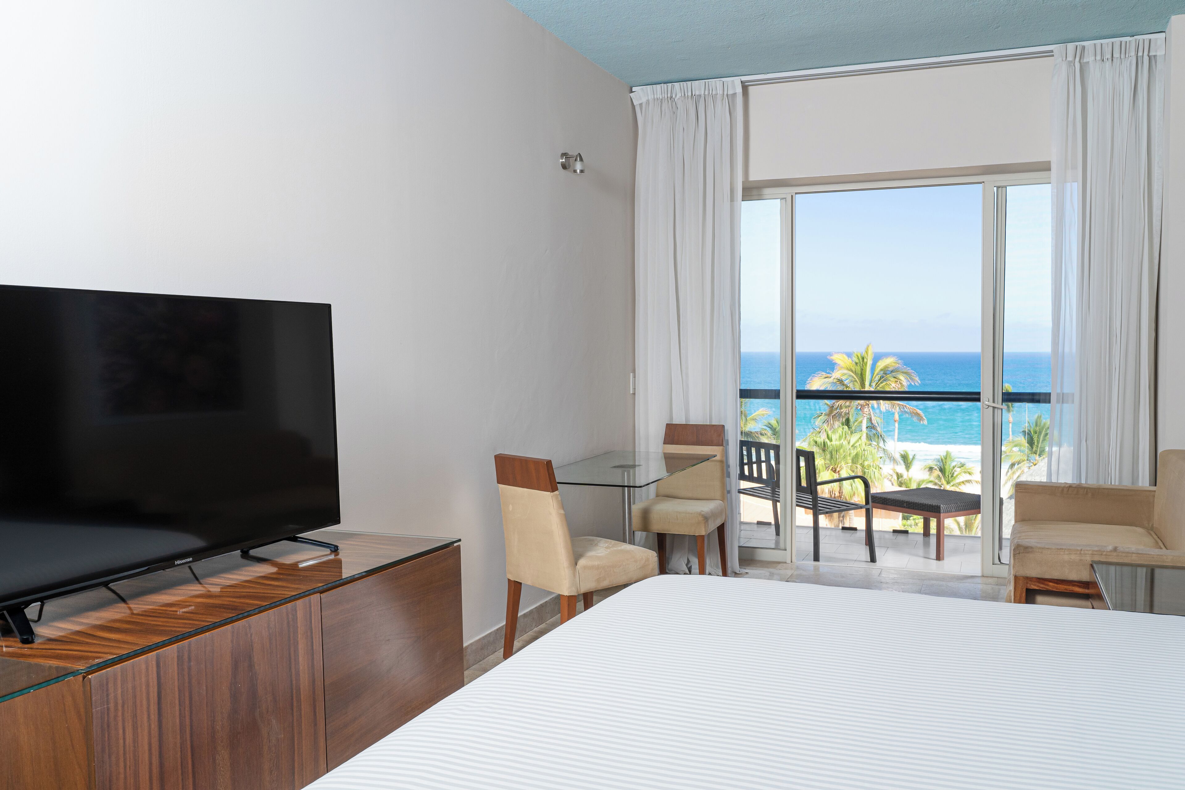 Habitación de hotel con cama, televisor, balcón con vista al océano y zona de comedor con sillas y mesa.