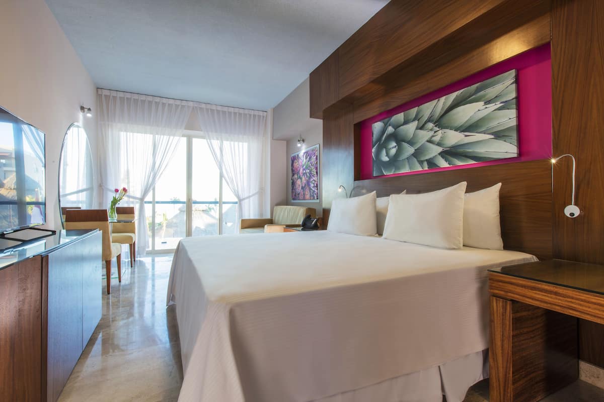 Ein Hotelzimmer mit einem großen Bett, einem Schreibtisch mit Computer, einem Sessel, einem Fenster mit Vorhängen und einem Bild an der Wand.