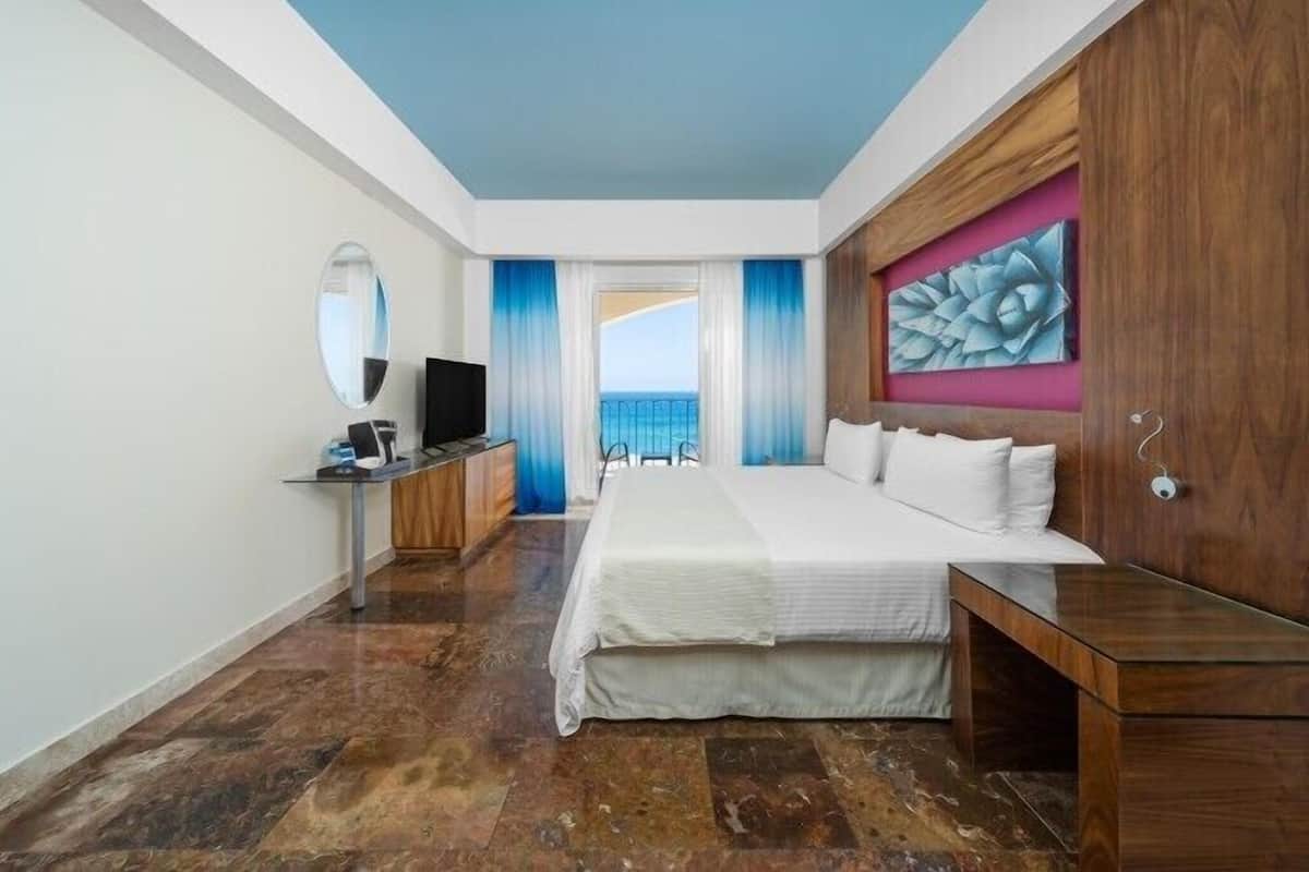 Ein Hotelzimmer mit einem großen Bett, einem Schreibtisch mit Computer, einem Fernseher und Blick aufs Meer durch ein Fenster.
