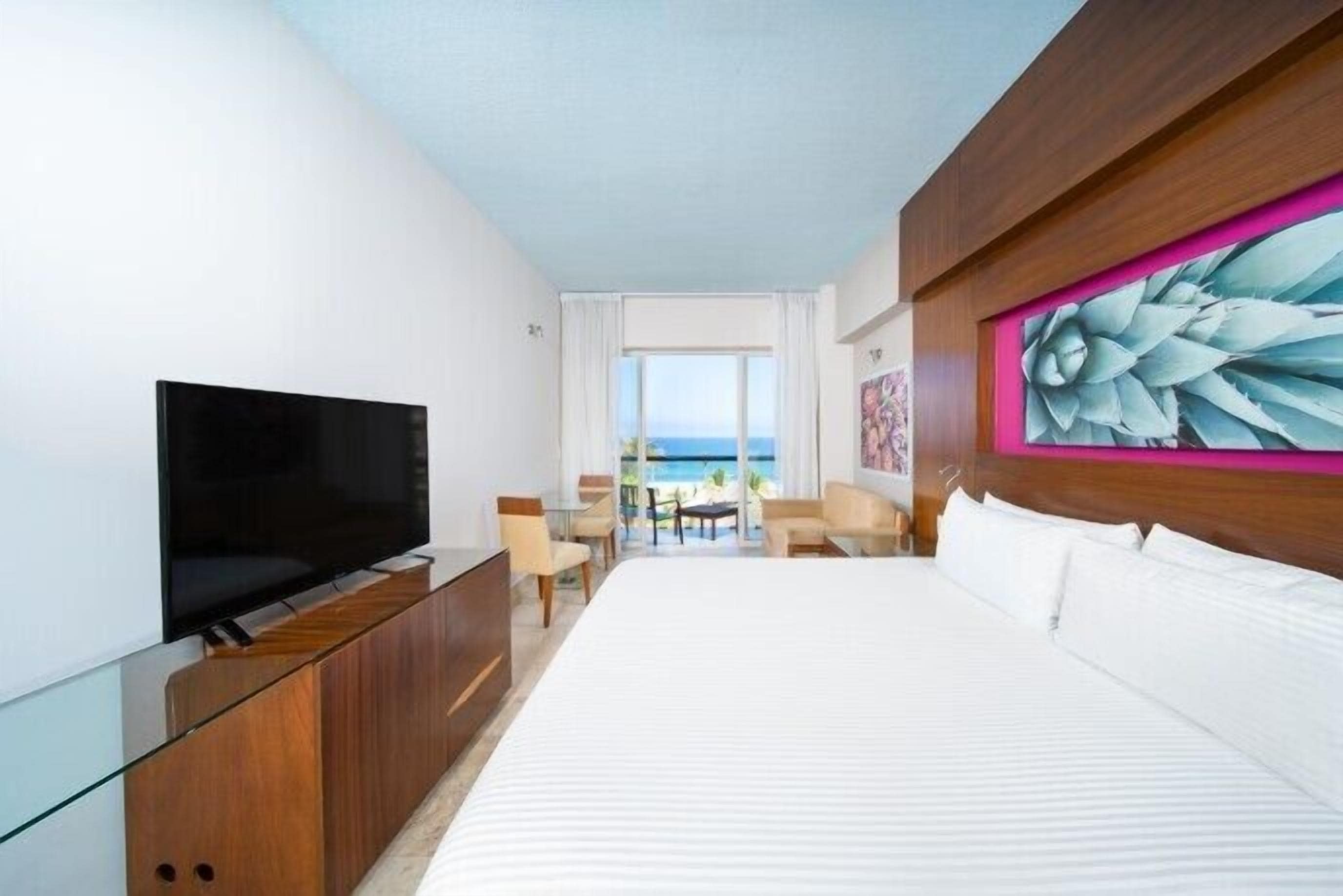A hotel room with a large bed, a TV on a wooden cabinet, a painting on the wall, and a view of the sea through the window.