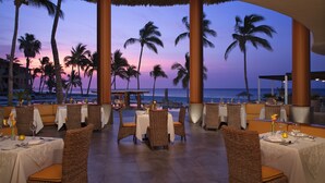 Un restaurant avec des chaises et des tables en osier, disposées pour le dîner, offrant une vue sur une plage bordée de palmiers et un coucher de soleil.