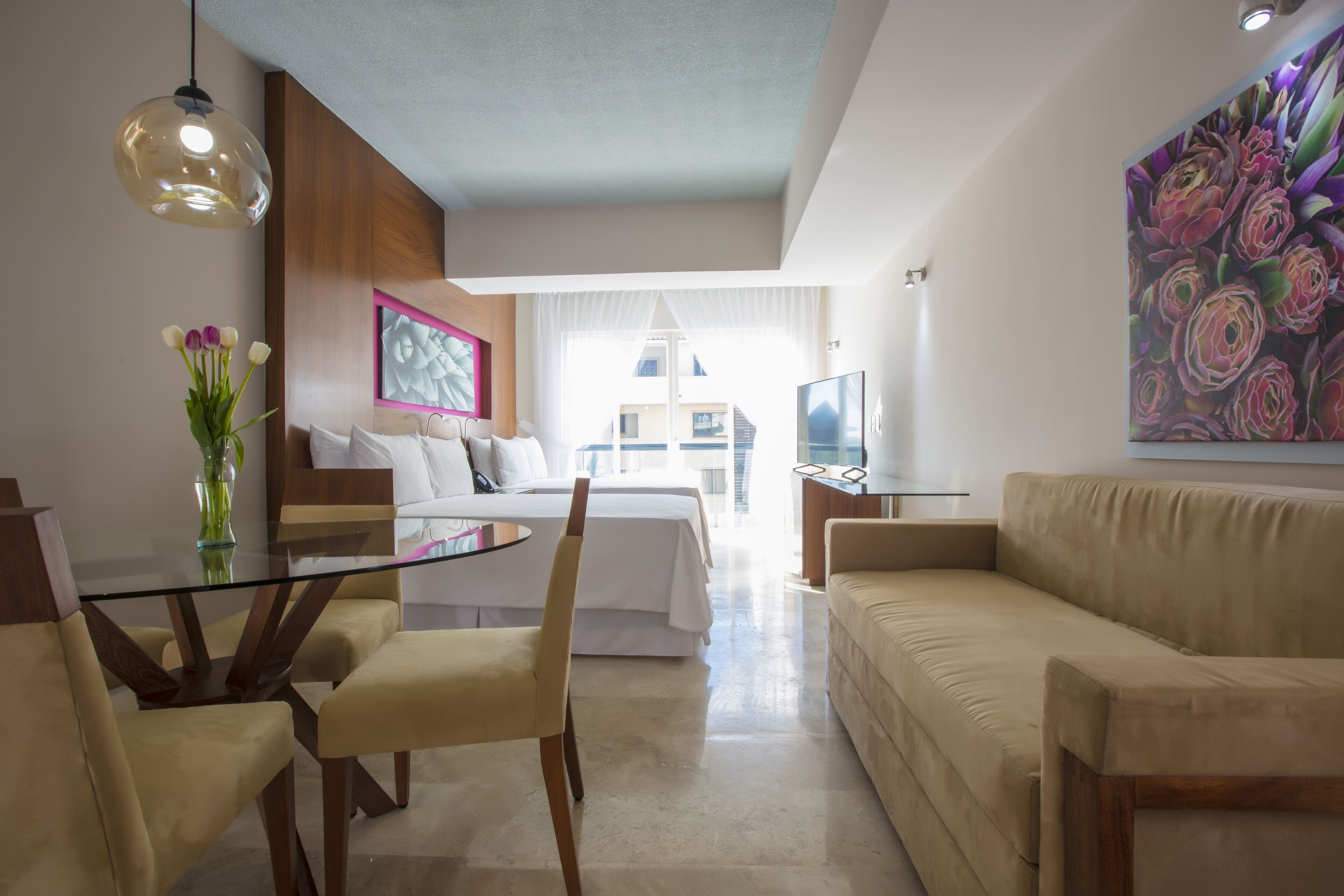 Une chambre d’hôtel moderne avec une table à manger en verre, un canapé beige et un grand tableau.