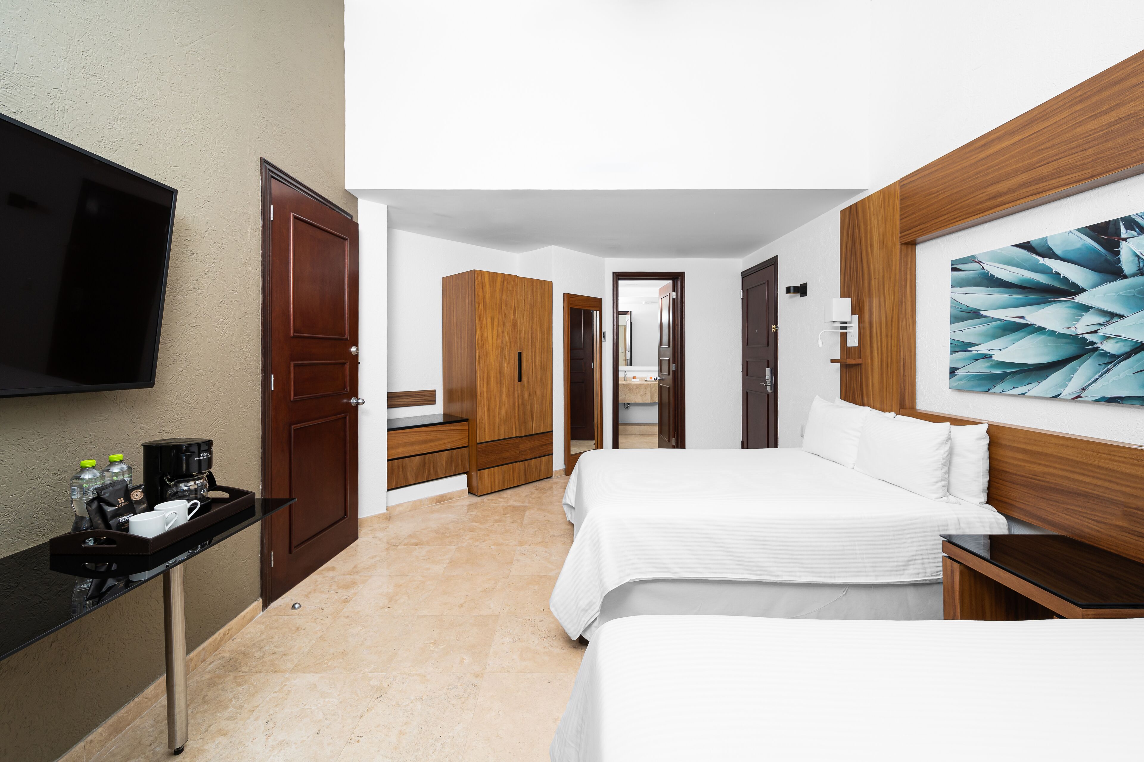A hotel room with a flat-screen TV, a wooden wardrobe, a bed with white bedding, a bedside table, and a large abstract painting on the wall.