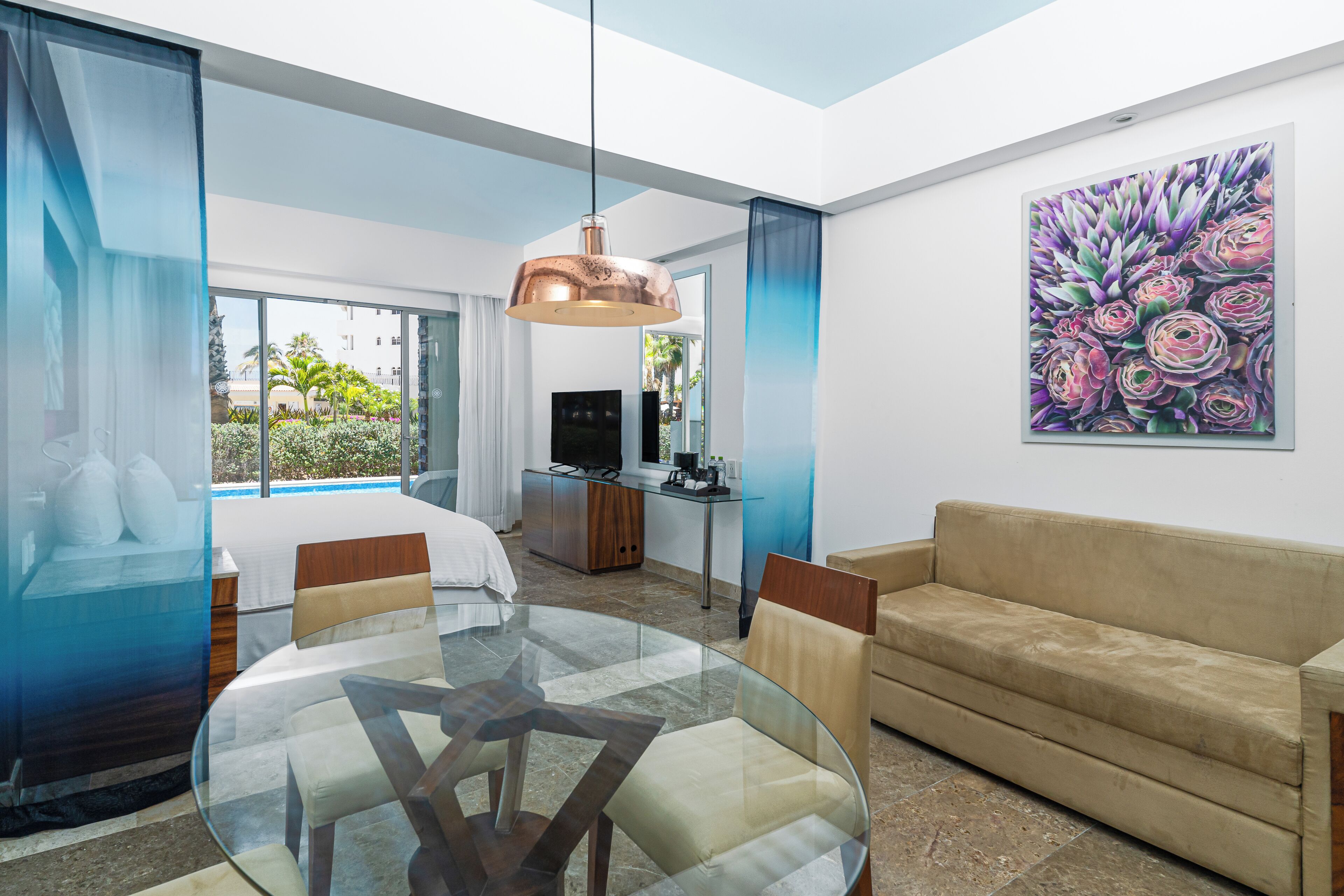 A modern hotel room with a glass dining table, a sofa, a TV, and a painting on the wall.