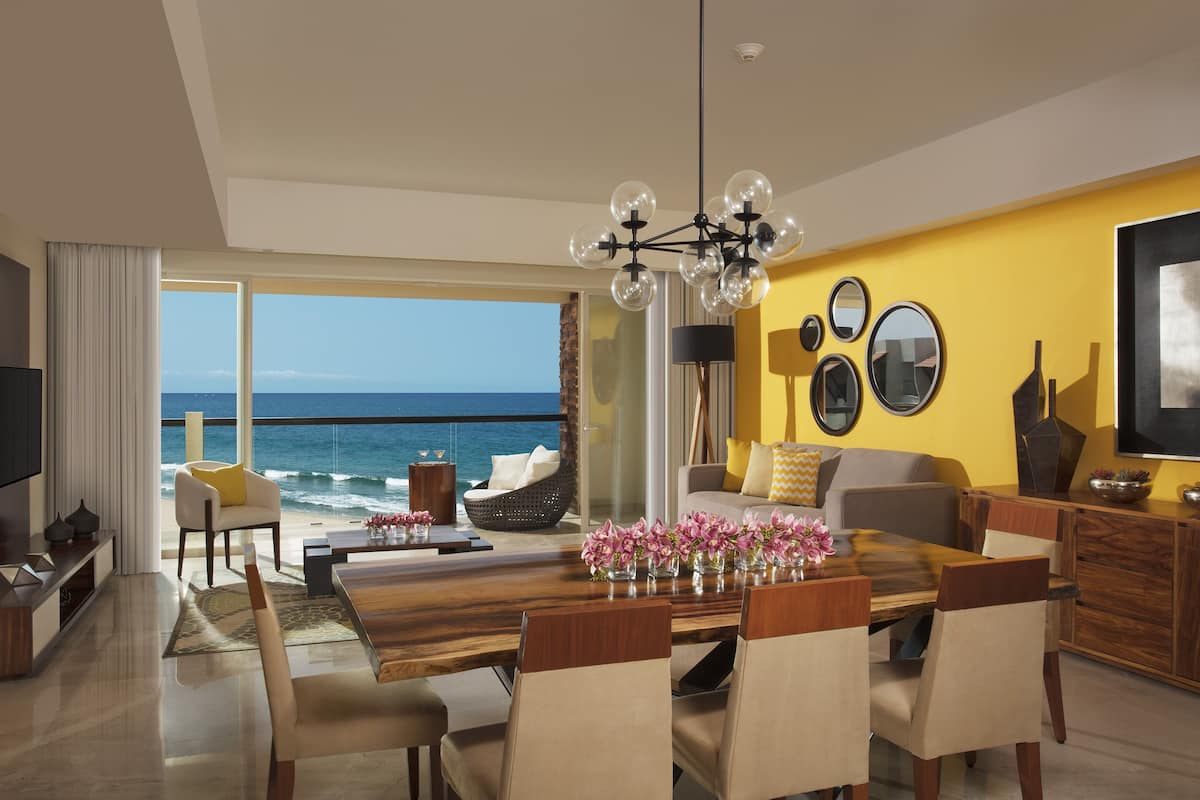 Ein modernes Wohnzimmer mit einem Holzesstisch, beigen Stühlen, einem Sofa mit gelben Kissen und einem großen Fenster mit Blick auf den Ozean.