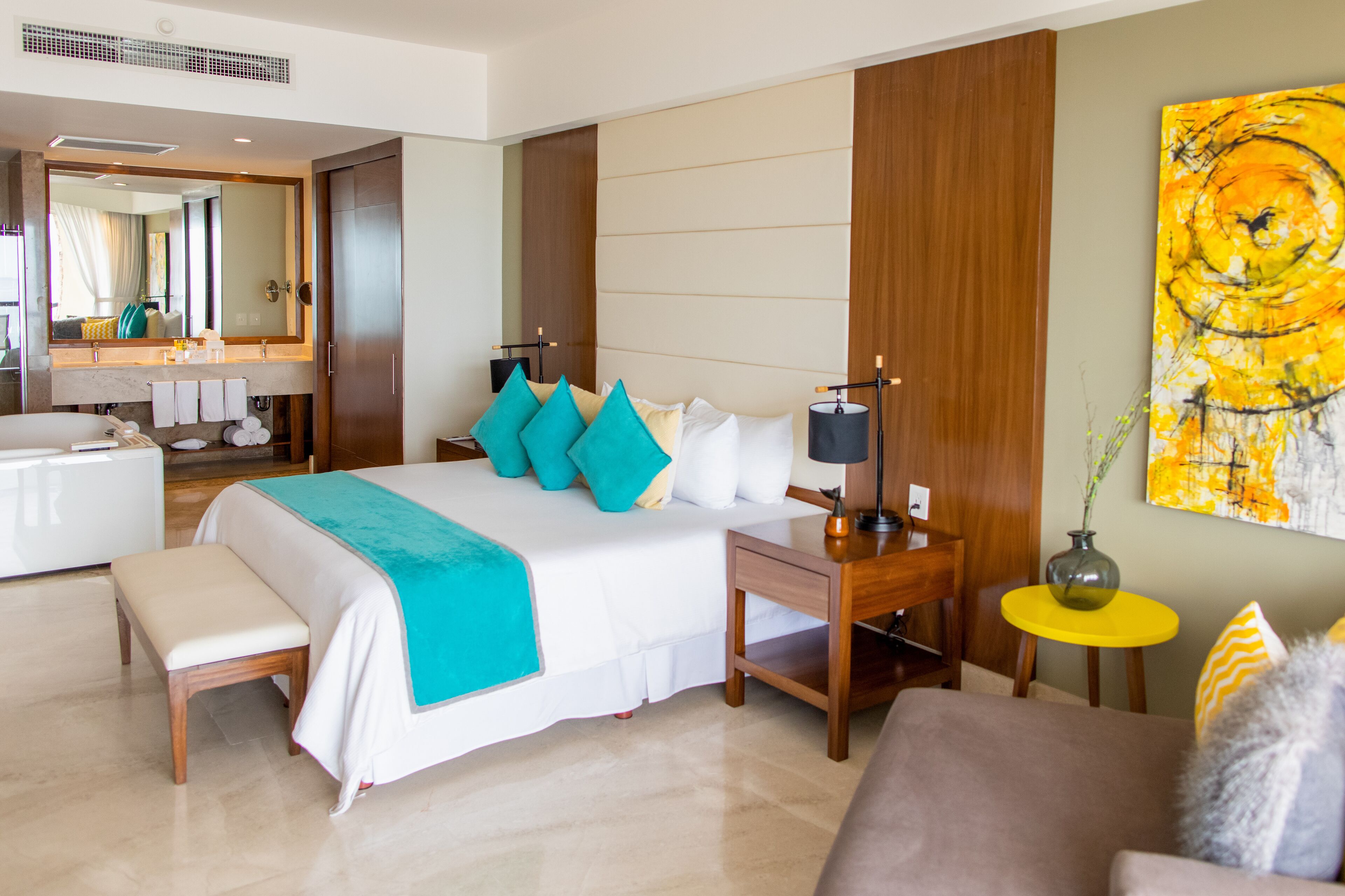 A modern hotel room with a large bed, bedside table, lamp, and a small round table with a vase.
