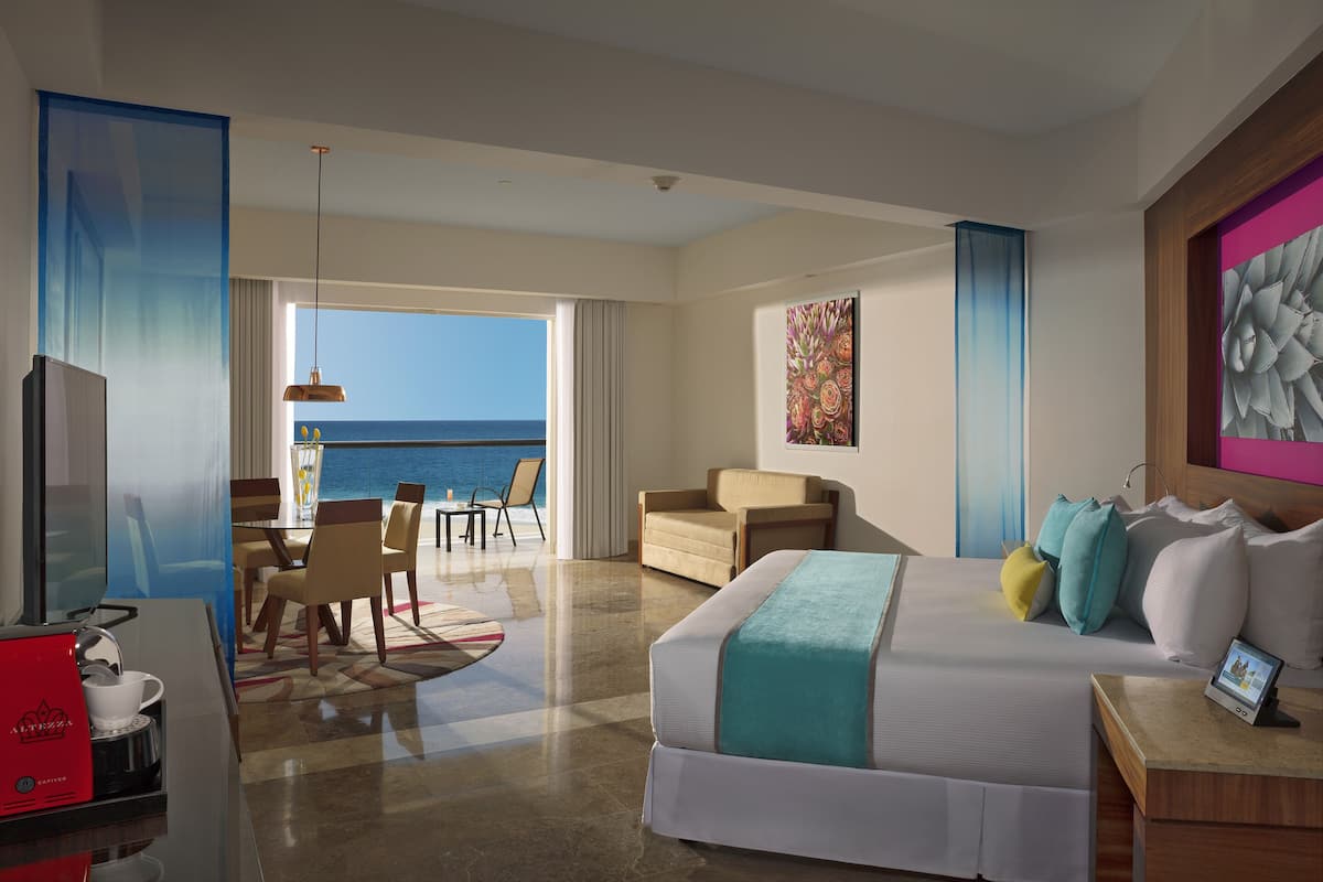 Ein Hotelzimmer mit Bett, Schreibtisch, Fernseher, Kaffeemaschine, Essbereich mit Stühlen und Tisch sowie Meerblick.