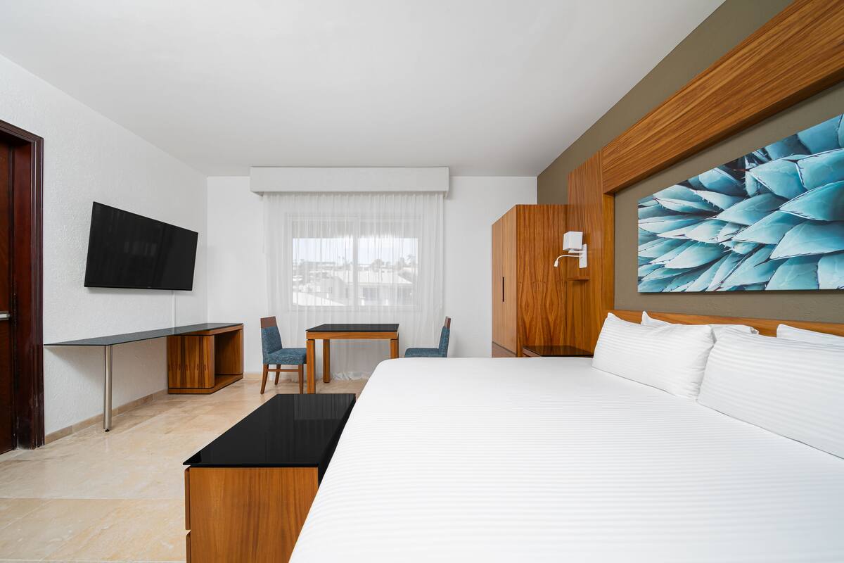 Ein Hotelzimmer mit einem großen Bett, einem Schreibtisch, einem Sessel, einem Fernseher und einem großen Fenster mit Vorhängen.