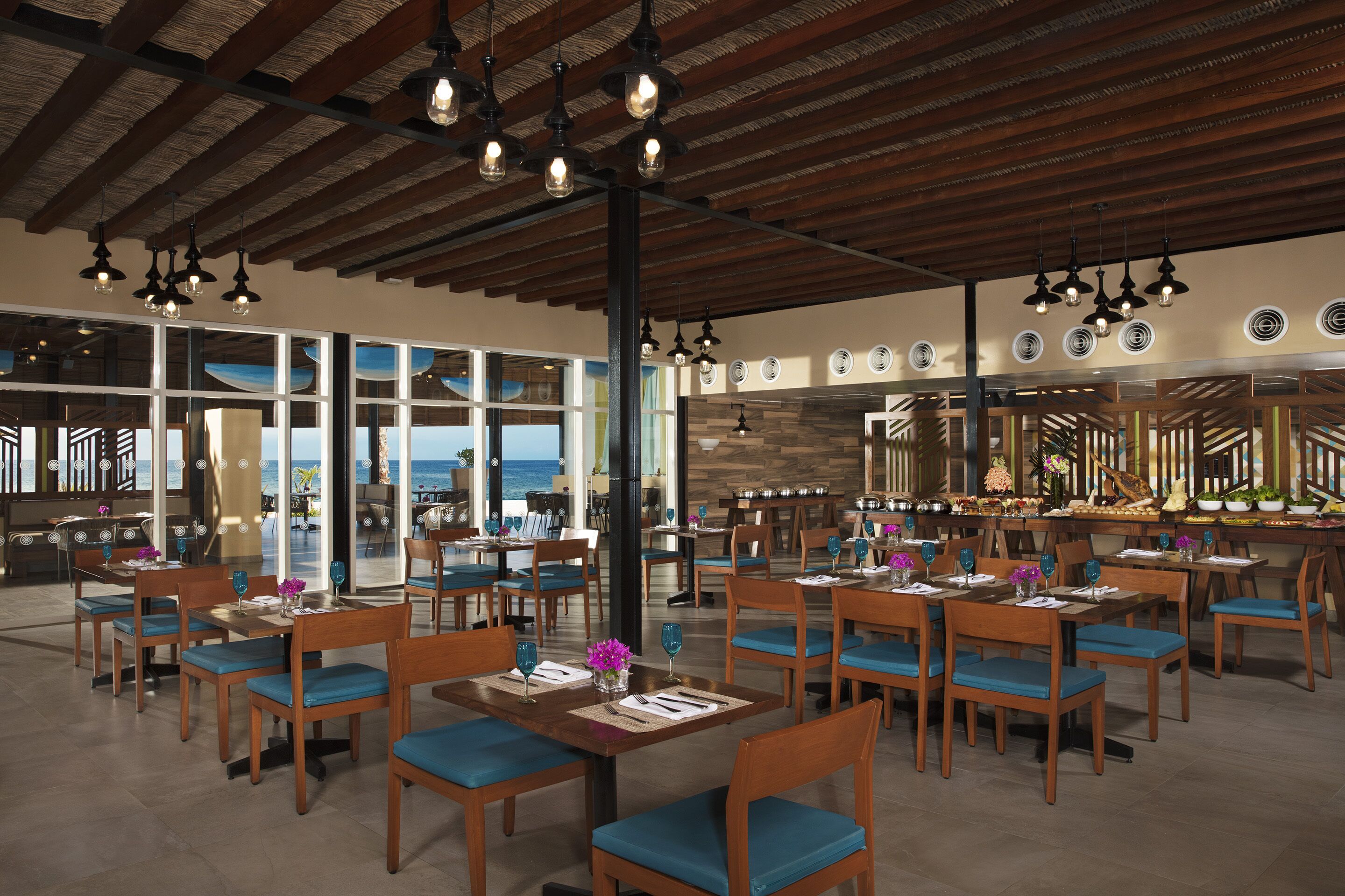 A spacious restaurant with wooden tables and chairs, large windows offering a view of the ocean, and a high ceiling with exposed beams.