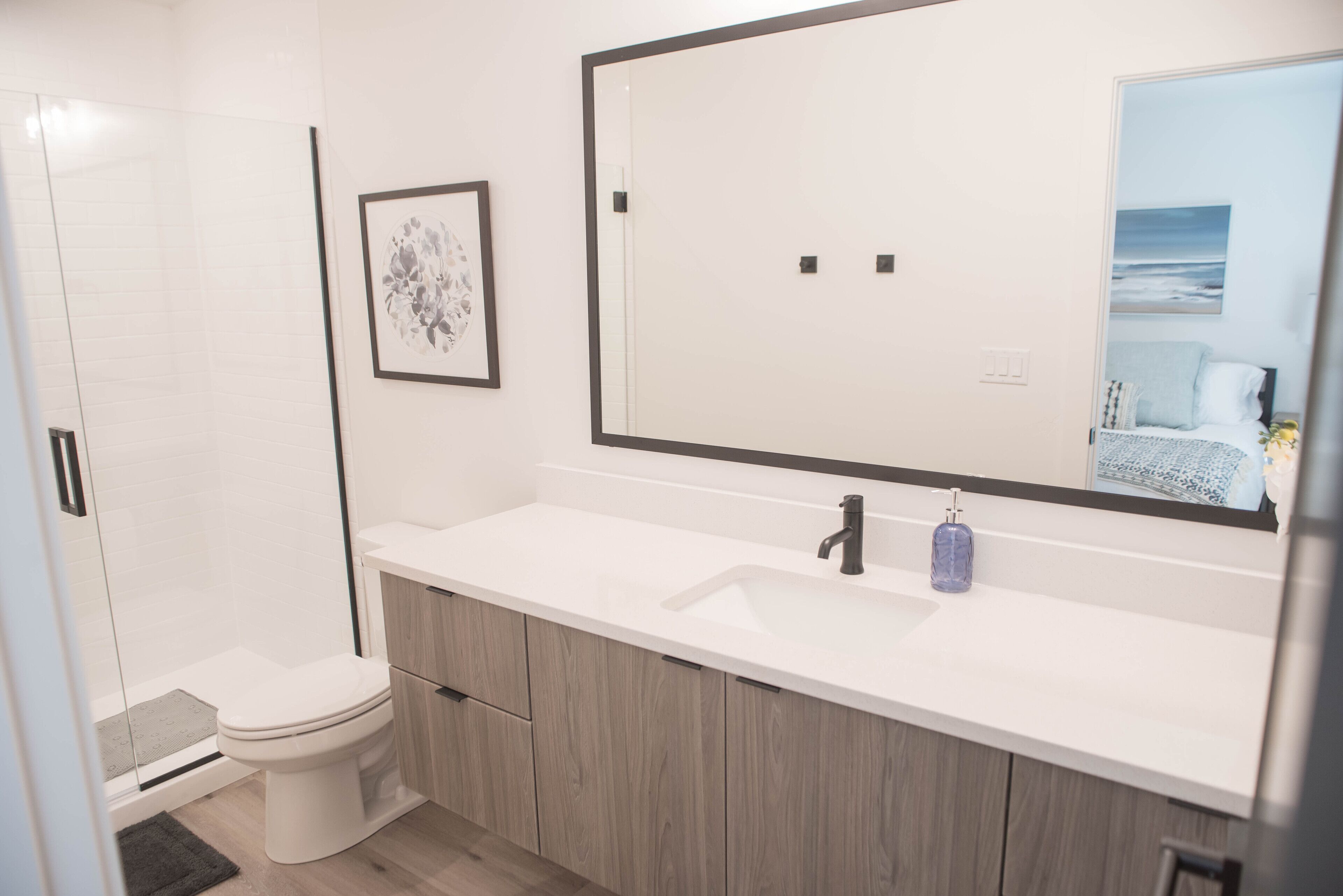 A modern bathroom with a white sink, wooden cabinets, a large mirror, and a framed artwork on the wall.