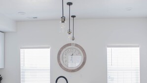 A modern kitchen with a white countertop, a sink, and a round clock on the wall. There are two windows with blinds and a ceiling light fixture.