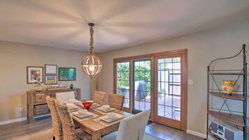 Un comedor con mesa de madera dispuesta para cuatro personas, sillas de mimbre, una lámpara colgante y vistas a un patio exterior a través de puertas de vidrio.