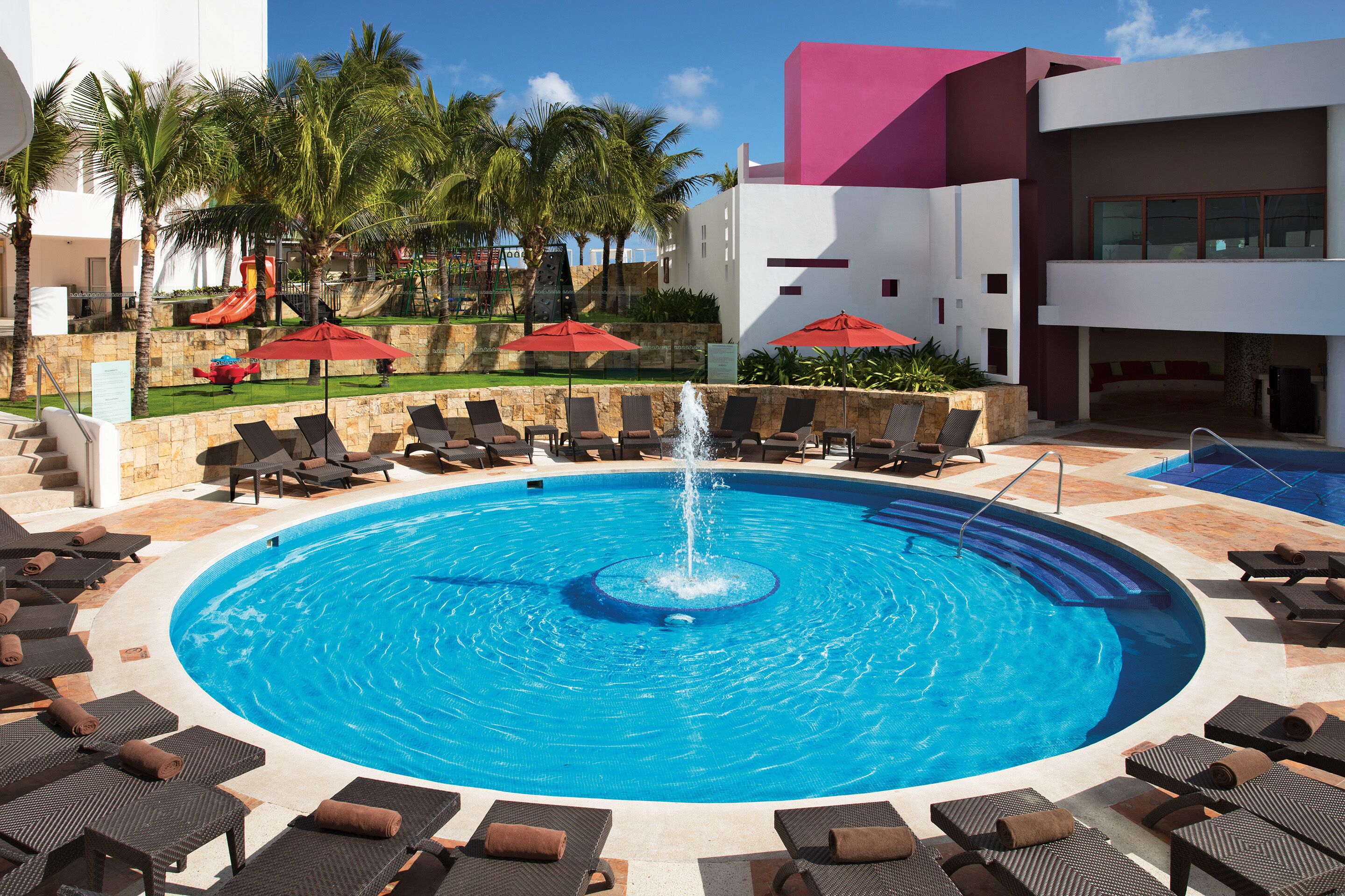 Una piscina con una fuente central, rodeada de tumbonas y sombrillas, con palmeras y un edificio moderno al fondo.