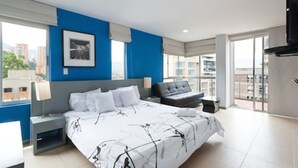 A modern hotel room with a large bed, a grey sofa, a small table, and a city view through large windows. - Loslan Real Estate (Medellín)