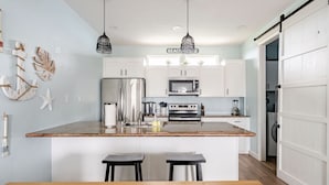 Une cuisine avec des meubles blancs, un réfrigérateur en acier inoxydable, un four à micro-ondes et une cuisinière. On y trouve un îlot central en bois avec deux tabourets, et des luminaires suspendus au-dessus.