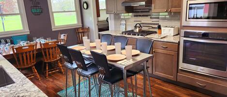 A modern kitchen with wooden cabinets, a stainless steel stove, and an oven. There is a dining area with a table set for four.