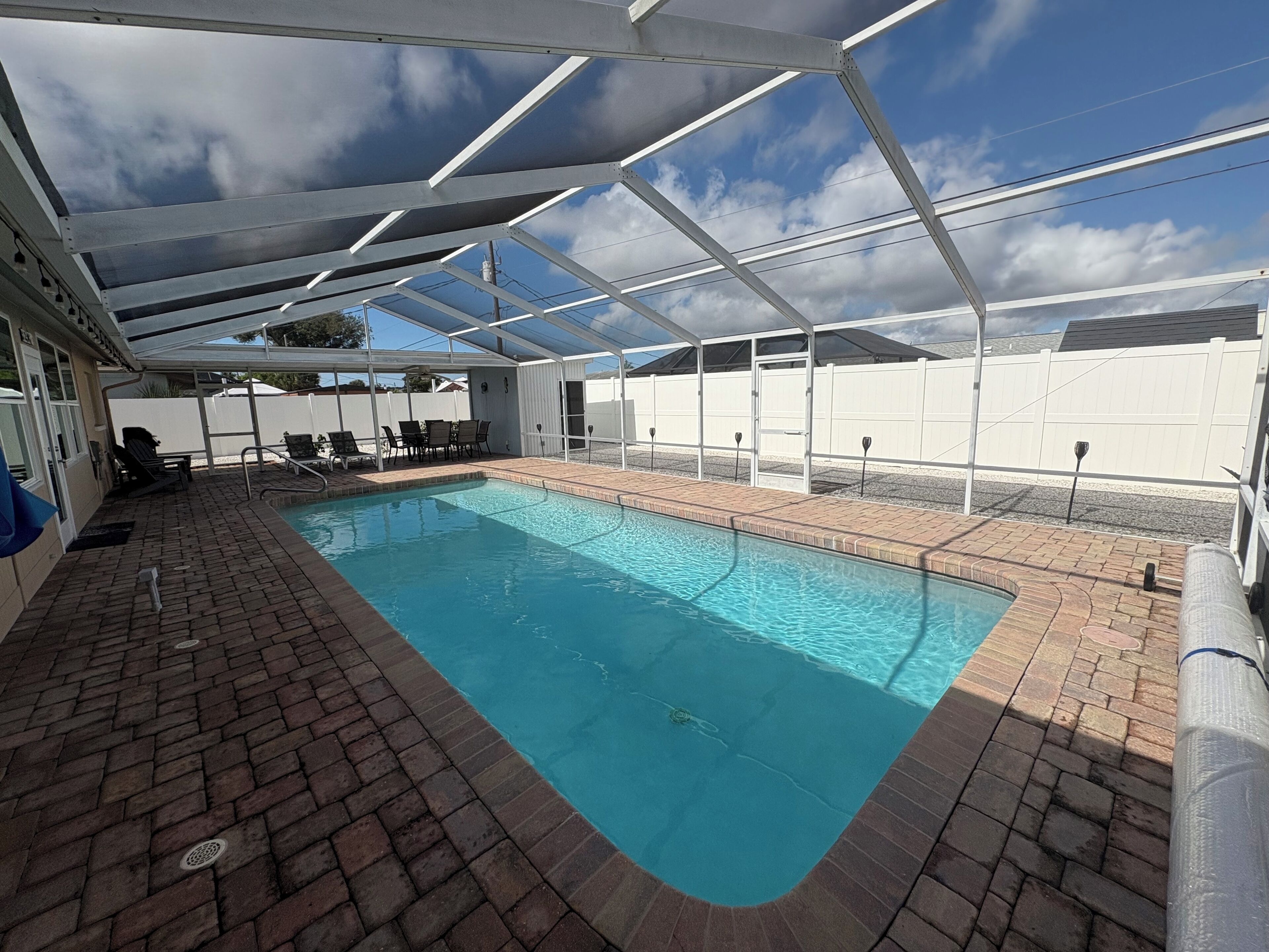 A covered swimming pool with a brick deck, lounge chairs, and a white fence in the background.