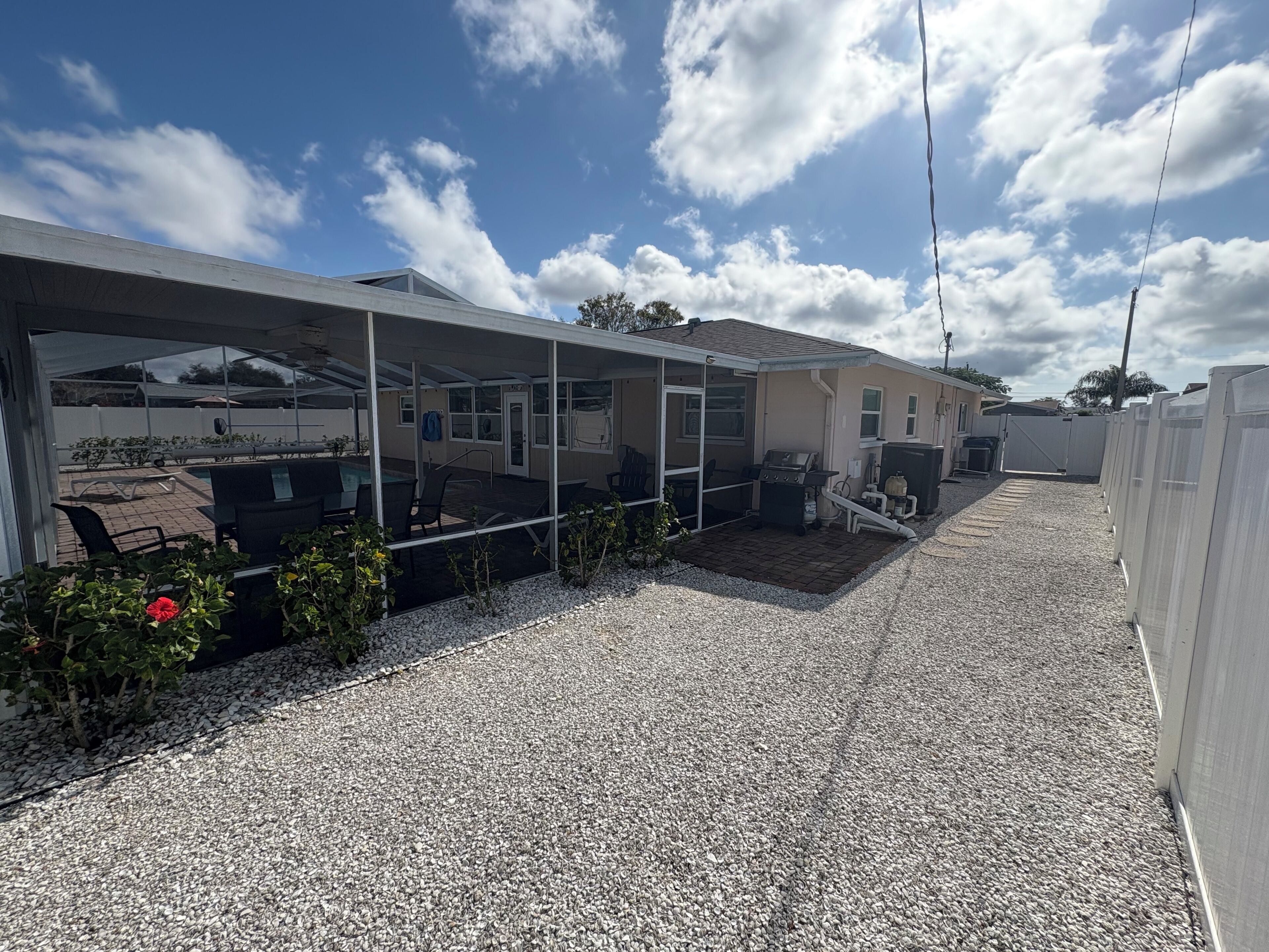 Un patio extérieur couvert avec un barbecue et un espace salon, une piscine et une allée en gravier.