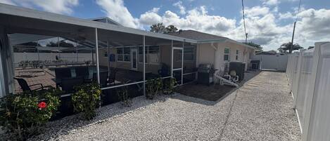 Un patio extérieur couvert avec un barbecue et un espace salon, une piscine et une allée en gravier.